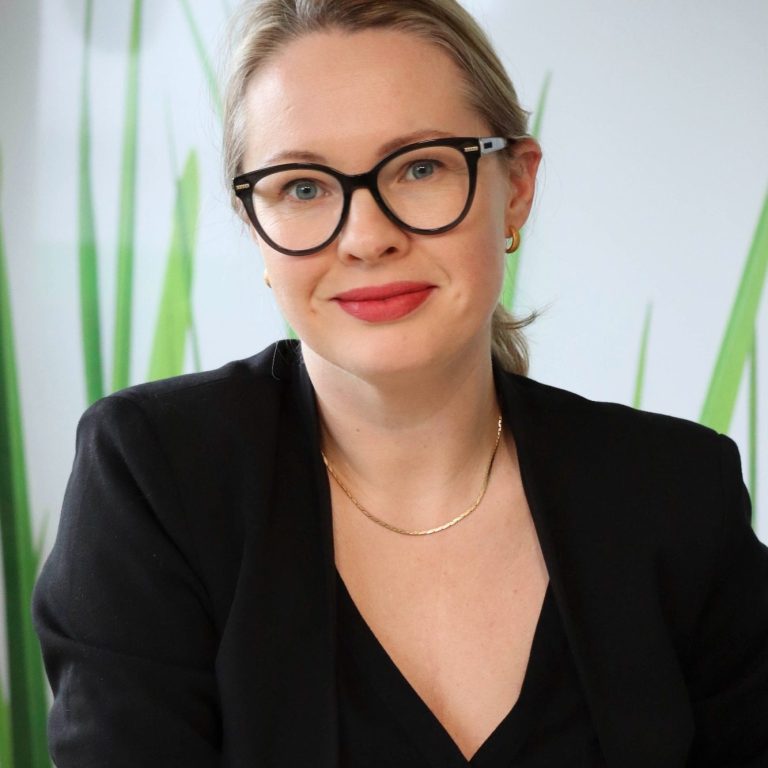 Frau mit blonden Haaren, Brille, schwarzem Blazer und neutralem Hintergrund mit grünen Pflanzen.